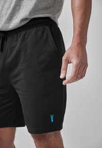 Pantaloni scurți negri din bumbac cu talie elastică, având buzunare laterale și un logo al homarului albastru pe piciorul stâng. Textură: netedă.