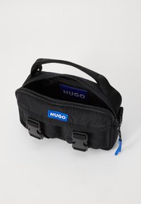 Black fabric bag with a top handle, zipper closure, and a blue HUGO logo. Features two black buckles and a textured surface.