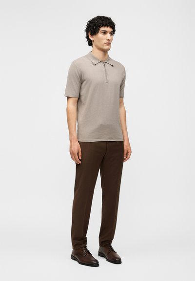 Jeune homme debout, portant un polo beige à manches courtes avec fermeture éclair, un pantalon marron et des chaussures habillées marron foncé sur un fond blanc uni.