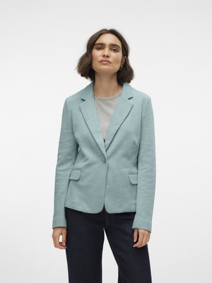 Donna con capelli corti e scuri che indossa un blazer azzurro chiaro sopra una maglietta grigia e pantaloni scuri, in piedi davanti a uno sfondo bianco uniforme.