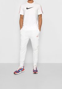 Bílý tričko Nike s černým logem, bílé tepláky s bočními kapsami a vícebarevné tenisky s červenými, modrými a bílými akcenty.