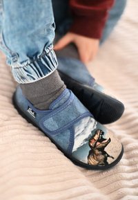 Kind trägt blaue Dinosaurier-Hausschuhe und graue Socken, sitzt mit einer Hand auf dem Bein und trägt hellblaue elastische Jeanshosen.