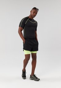 Maglietta atletica nera con accenti verdi, abbinata a pantaloni corti neri sopra pantaloni a compressione neon verdi. Scarpe da corsa nere completano l'outfit.