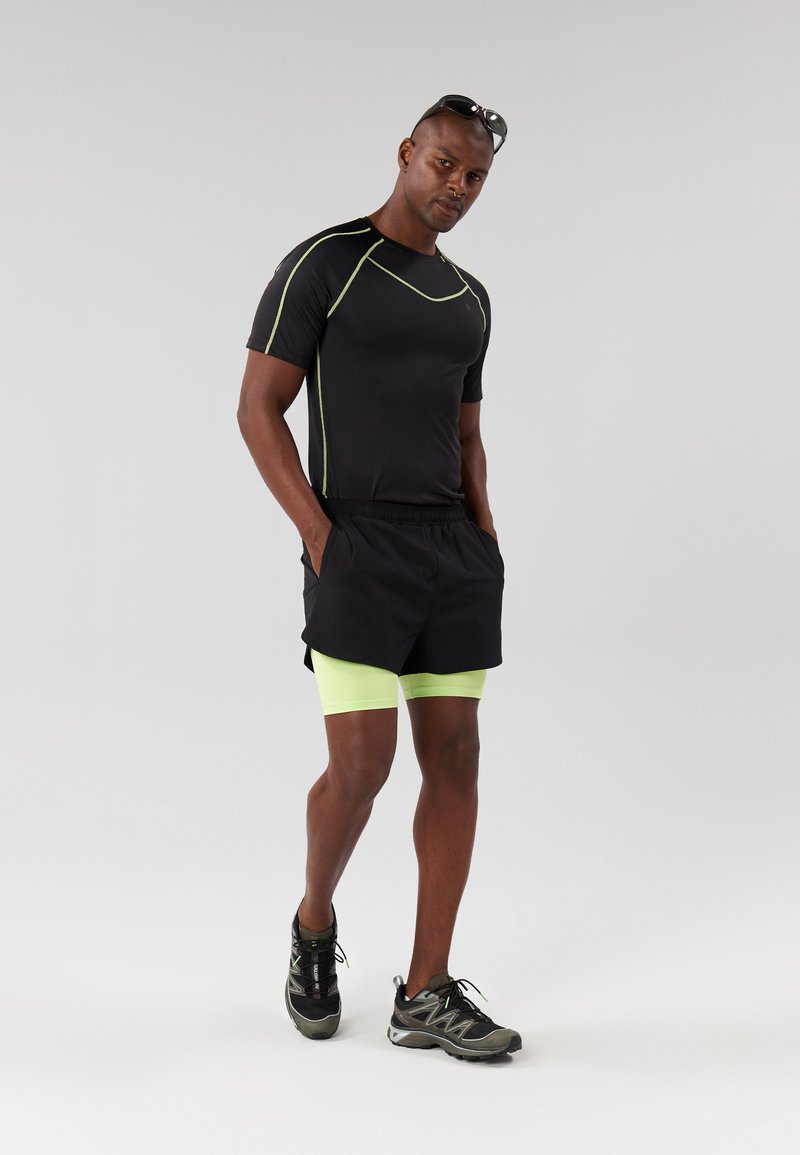 Camisa atlética negra con detalles en verde, combinada con pantalones cortos negros sobre pantalones cortos de compresión verde neón. Zapatillas de correr negras completan el conjunto.