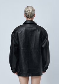 Veste surdimensionnée en cuir noir avec un col à revers, une finition texturée, des coutures latérales et un patch logo discret à l'arrière.
