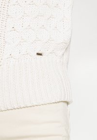 Vit stickad tröja med en strukturerad mönster och ribbad fåll. Har en liten metallisk logotagg på sidan.