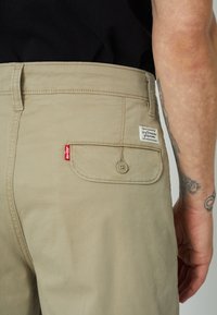 Pantalones cortos de algodón color beige con un bolsillo trasero y cierre de botón. Presentan una etiqueta roja de Levi's y una etiqueta blanca cosida en el bolsillo.