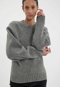 Pull en tricot gris avec un col rond, épaules tombantes et ourlet côtelé. Texture douce, coupe oversize, et manches longues et larges.