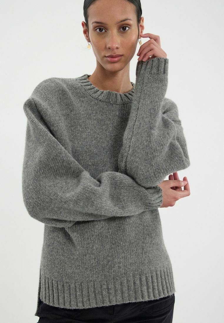 Pull en tricot gris avec un col rond, épaules tombantes et ourlet côtelé. Texture douce, coupe oversize, et manches longues et larges.