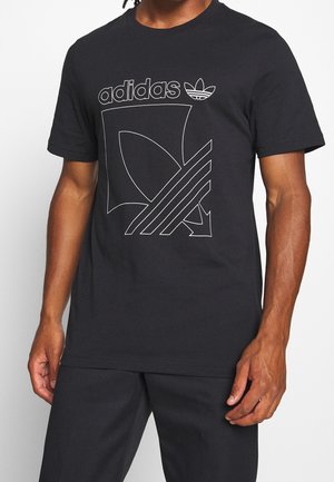 Homme portant un T-shirt noir Adidas avec un grand logo tréfle en contour et un design de texte sur le devant, assorti à un pantalon noir.