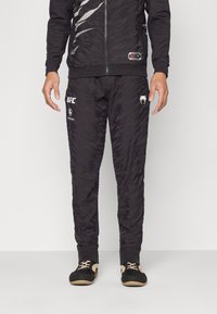 Sort atletisk tracksuit som har en tekstureret, mønstret jakke og bukser. Inkluderer UFC-branding på venstre ben samt en lynlåsåbning.