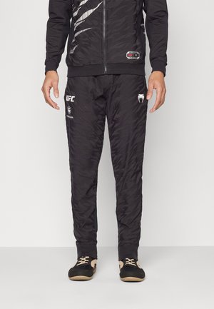 Chándal deportivo negro con una chaqueta y pantalones texturizados y con patrón. Incluye la marca de UFC en la pierna izquierda y un cierre de cremallera.