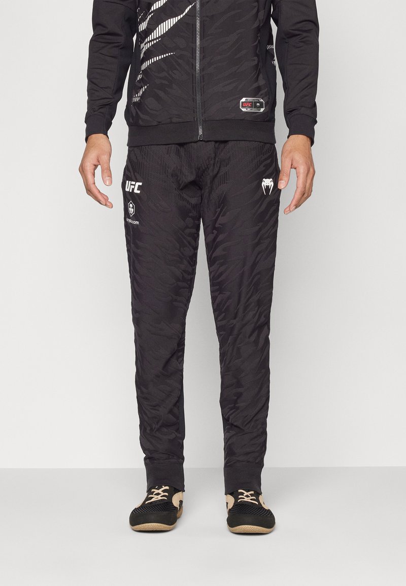 Sort atletisk tracksuit som har en tekstureret, mønstret jakke og bukser. Inkluderer UFC-branding på venstre ben samt en lynlåsåbning.