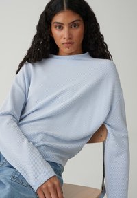 Hellblauer Strickpullover mit weitem Schnitt, versehen mit einem strukturierten Muster und weiten Ärmeln, kombiniert mit hellen Jeans.