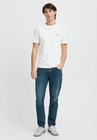 Weißes Baumwoll-T-Shirt mit einem kleinen grünen Fahrradgrafik auf der Brust, kombiniert mit blauen Jeans und grauen Sneakers.