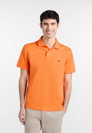 Uśmiechnięty młody mężczyzna o krótkich ciemnych włosach, ubrany w pomarańczową koszulkę polo i jasnobeżowe spodnie, stojący z jedną ręką w kieszeni.