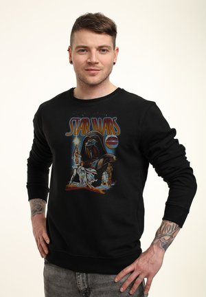 Junger Mann mit Tattoos trägt einen schwarzen Sweatshirt mit Retro Star Wars-Figuren und Logo, steht mit den Händen in der Hüfte.