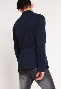 Giacca casual blu navy con un design strutturato, toppe ai gomiti e una texture liscia, abbinata a jeans grigi attillati.