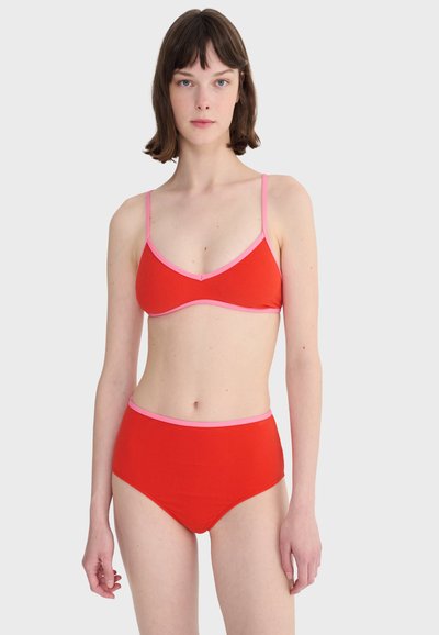 Femme debout, face à l'avant, portant un bikini rouge avec des bordures roses sur un fond gris clair uni.