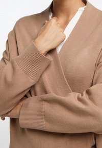Cardigan lavorato a maglia marrone chiaro con design avvolgente, polsini a coste e texture morbida, indossato su un top bianco con scollatura a V.