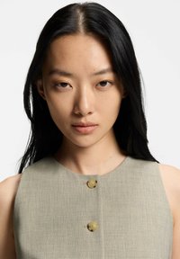 Bezrękawnik khaki z okrągłym dekoltem, ozdobiony dwoma dużymi, jasnymi guzikami z przodu oraz teksturowanym wykończeniem materiału.