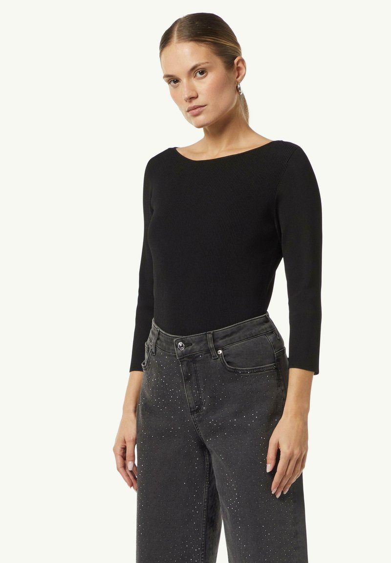 Schwarzes, geripptes Langarmoberteil mit weiter Rundhalsöffnung, kombiniert mit hochtaillierten schwarzen Jeans, die an der Vorderseite metallische Nieten haben.