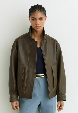 Junge Frau mit geflochtenem Haar trägt eine übergroße dunkelbraune Lederjacke, ein marineblaues Top, hellblaue Hose und einen schwarzen Gürtel mit goldener Schnalle.
