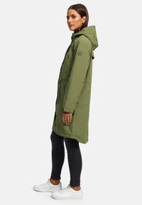 Olivengroen gewatteerde parka met ritszakken, verstelbare zoom en logo-detail op de mouw. Draagt over donkere jeans en witte sneakers.