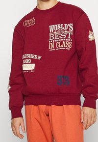 Felpa color bordeaux con varie scritte in crema e blu, con collo e polsini a coste, abbinata a pantaloni arancioni.