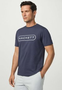 Námořnická modrá bavlněná tričko s krátkým rukávem a kulatým výstřihem. Na přední straně se nachází bílé logo "HACKETT" v oválném tvaru.