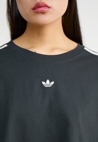 Melnas kokvilnas t-krekls ar apaļu apkakli, kas dekorēts ar baltu Adidas logo un baltām svītrām uz piedurknēm. Gluda tekstūra.