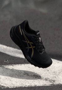 Black running shoe with a textured mesh upper, gold accent stripes, padded collar, and rubber sole. Features perforations for breathability.