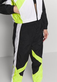 Färgglad träningsoverall med svarta, vita och neongröna paneler. Tyget verkar lätt och har en glänsande yta samt elastiska muddar.