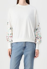 Sweat-shirt blanc avec manches ballon à motif floral, poignets et ourlet côtelés. Porté avec un jean bleu clair. Matière en coton doux.