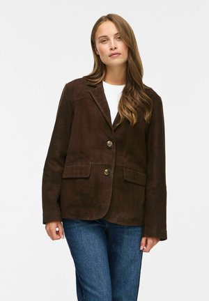 Donna con capelli lunghi e castani che indossa una giacca marrone scuro abbottonata, camicia bianca e jeans blu, in piedi davanti a uno sfondo semplice.