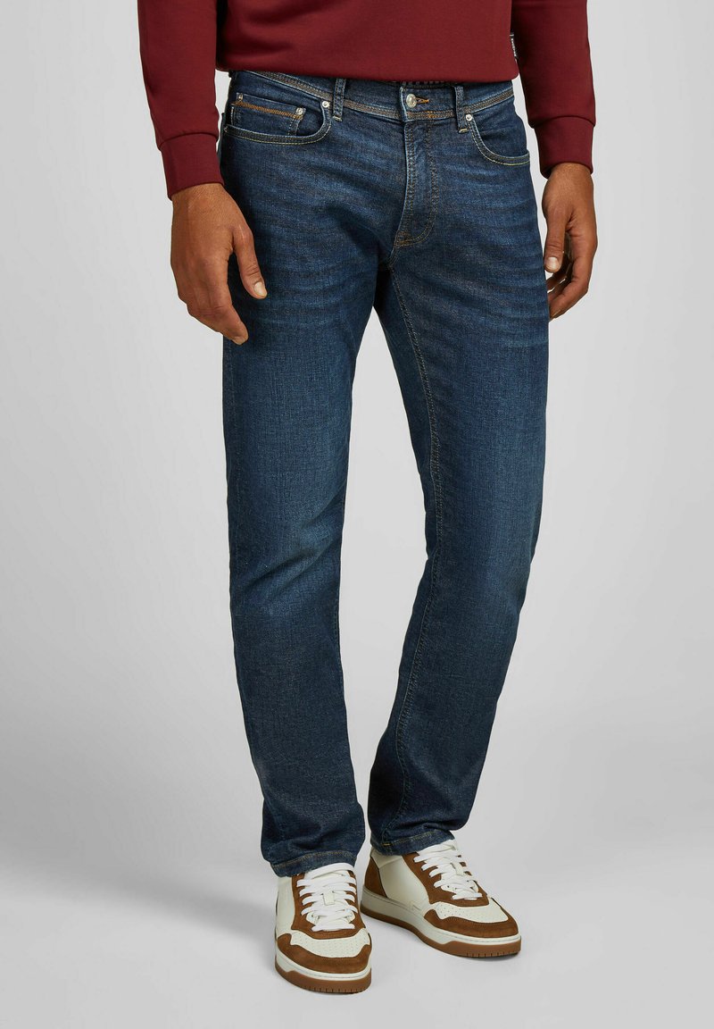 Jean en denim bleu avec une coupe slim, dotés d'un design à cinq poches, d'une légère décoloration et de coutures en cuivre, associés à des baskets blanches et brunes.