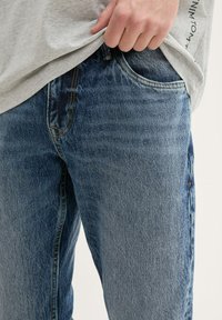 Persona con jeans de mezclilla azules y una camisa gris claro, con una mano parcialmente metida en el bolsillo delantero de los jeans.