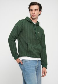 Hanorac verde cu modelă în formă de romb, cu glugă cu șnur, buzunar frontal și manșete cu nervuri. Purtați peste o cămașă albă, asortat cu blugi de culoare albastru deschis.