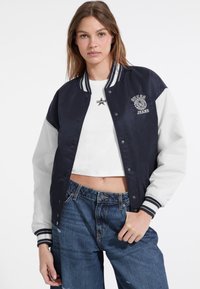 Marineblauwe en witte kleurgeblokte bomberjack met drukknoopsluitingen, ribgebreide manchetten en geborduurd logo. Gemaakt over een witte cropped shirt en denim jeans.