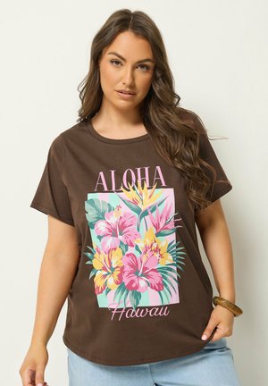 Frau trägt braunes T-Shirt mit bunten tropischen Blumen und dem Schriftzug "ALOHA Hawaii", kombiniert mit hellblauen Jeans und einem Holzarmband.