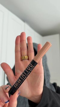 Matita occhi colorata per tatuaggi in un tubo nero con scritte bianche audaci, con tappo beige. Mano con anello d'oro che tiene la matita.