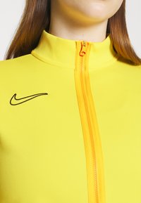 Yellow zip-up sports jacket made of smooth fabric, featuring an orange zipper and a black Nike logo on the left chest area.