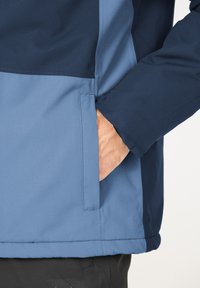Vandafvisende jakke i blå og marine, med en sidelomme til hænderne, kontrasterende teksturer og en glat overflade på stoffet.