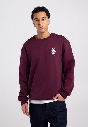 Bordeauxfarbener Sweatshirt mit Rundhalsausschnitt, gerippten Bündchen und Saum, mit einer kleinen, cremefarbenen Stickerei auf der Brust. Weicher Stoff und entspannte Passform.