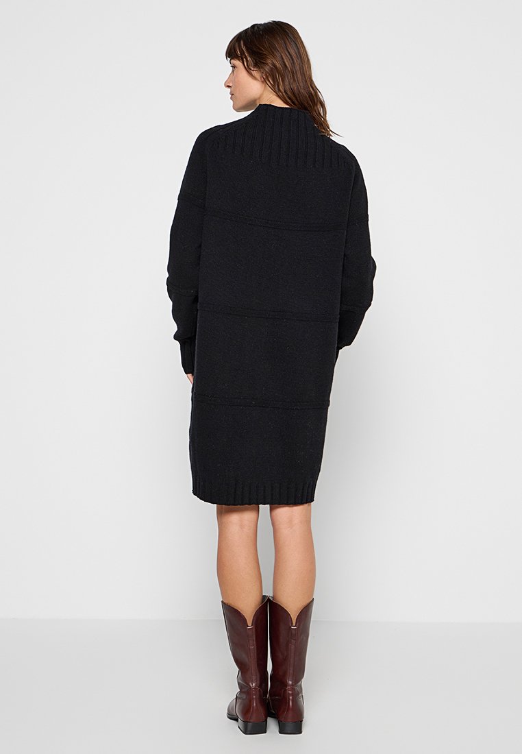Schwarzes Strickkleid mit geribbeltem Kragen und Bündchen, in kastiger Form und mit paneelartiger Textur. Abgerundet mit braunen knielangen Stiefeln.