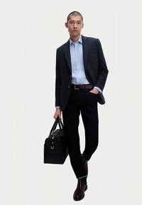 Herrenoutfit mit einem dunklen, strukturierten Blazer, hellblauem Hemd, dunklen Jeans, braunen Lederstiefeln und einer schwarzen Ledertasche mit silberner Hardware.