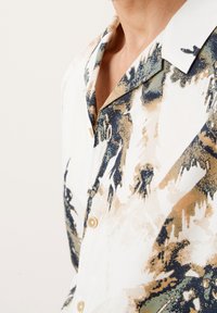 Camisa de manga corta en tela ligera con base crema y patrones abstractos en verde, azul y marrón; cuenta con cuello y cierre de botones.