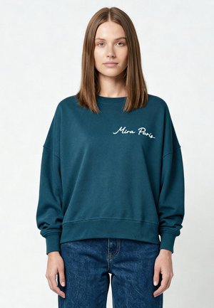 Jonge vrouw met stijl bruin haar die een teal sweater met ronde hals en de tekst "Mira Paris" draagt en blauwe spijkerbroek, staand tegen een witte achtergrond.