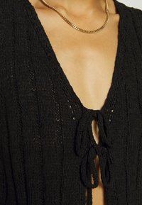 Black knitted garment featuring a deep V-neck, tied closure, and textured pattern. Accented with a gold chain necklace.