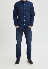 Muž v tmavě modré košili na knoflíky, tmavě modrých džínách a černých teniskách s bílými podrážkami, stojící s rukama v kapsách.